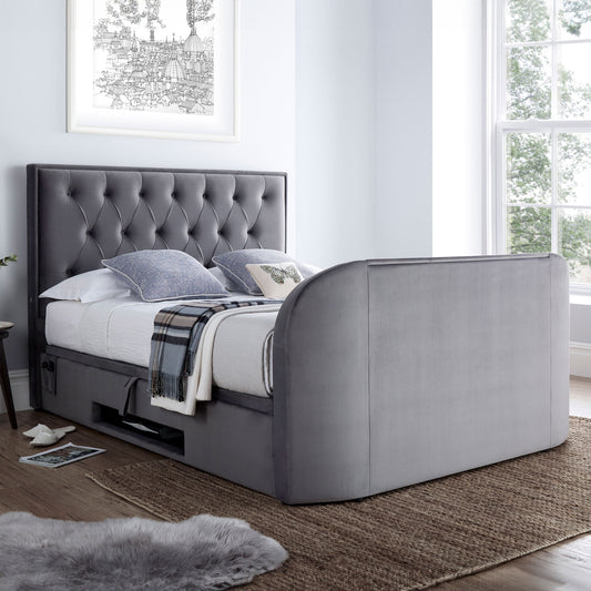 A grey velvet Lyon Ottoman TV Bed with a 2.1 media capacity and a 43" TV platform, featuring a tufted headboard and storage compartment beneath the bed.